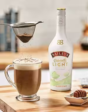 Bottle of Baileys Deliciously Light next to frothy coffee being sprinkled with chocolate powder