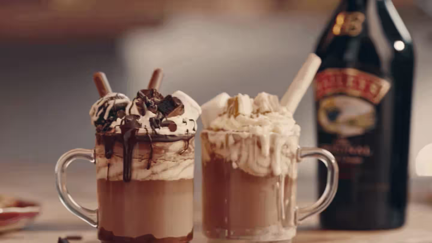 Two hot chocolates in glass mugs, topped with whipped cream and chocolate drizzle, with a Baileys bottle in the background.