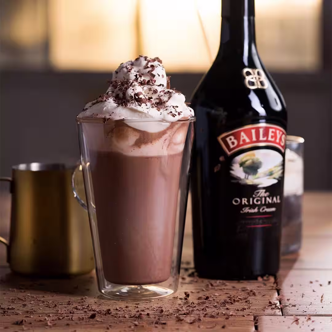 There’s a Baileys hot chocolate in a tall glass, next to a Baileys bottle. It's smooth and topped with whipped cream and chocolate.