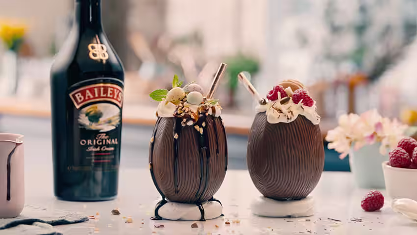 Two chocolate Easter egg cocktails filled with Baileys, topped with whipped cream, berries, and mini eggs, with a Baileys bottle in the background.