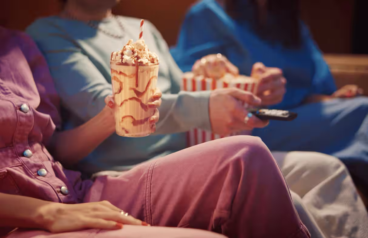 Three friends enjoy a movie night with a caramel-drizzled milkshake topped with popcorn, striped straws, and classic popcorn buckets.
